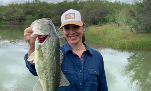 Smiling woman displaying a large fish caught by a peaceful pond surrounded by vegetation at El Estribo Ranch.